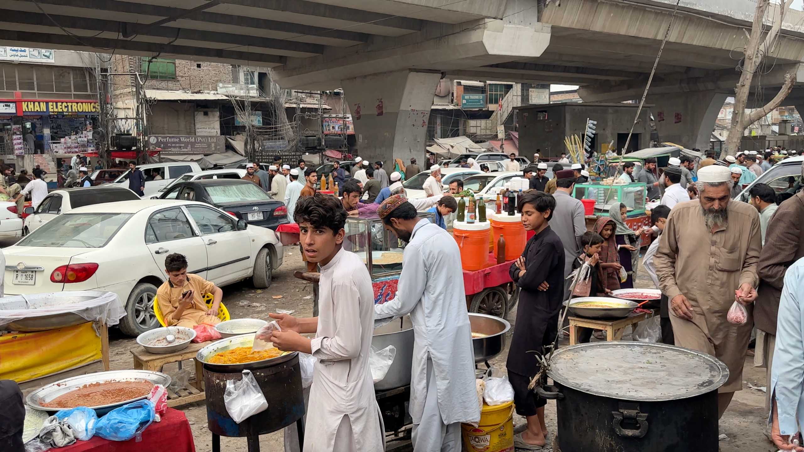 Bustling street food stall under a bridge, with crowds of people and busy traffic.