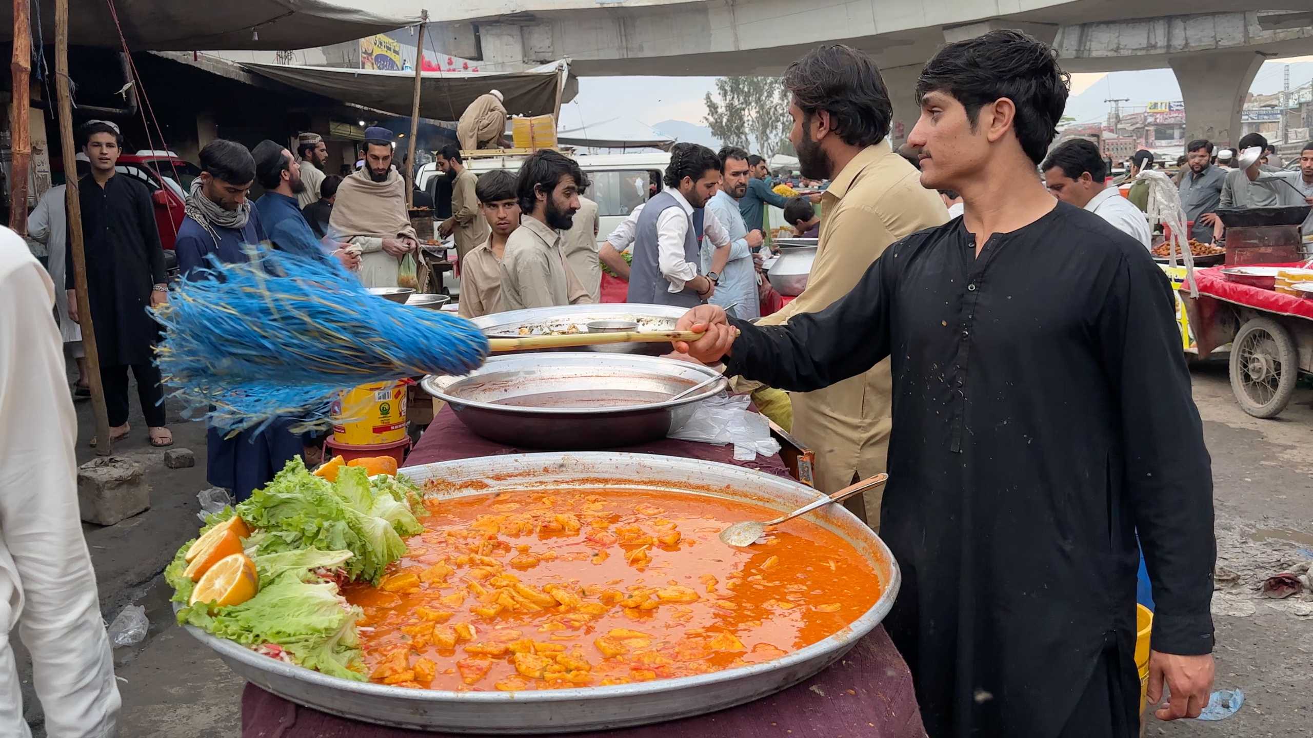 Young man stirring a large pot of curry, surrounded by hungry customers.