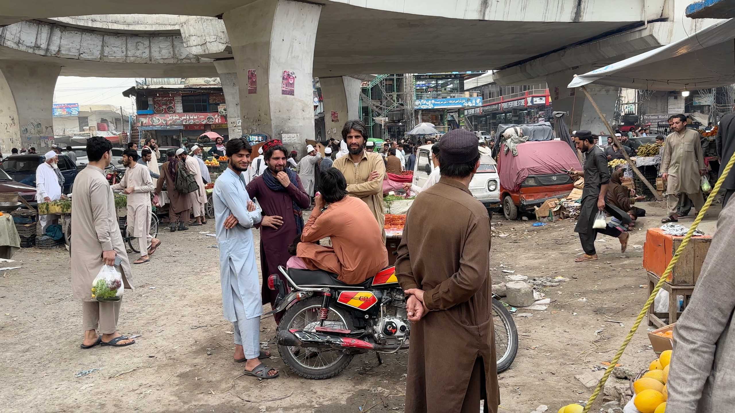 Groups of people engaged in conversations under the flyover, market life bustling around them.