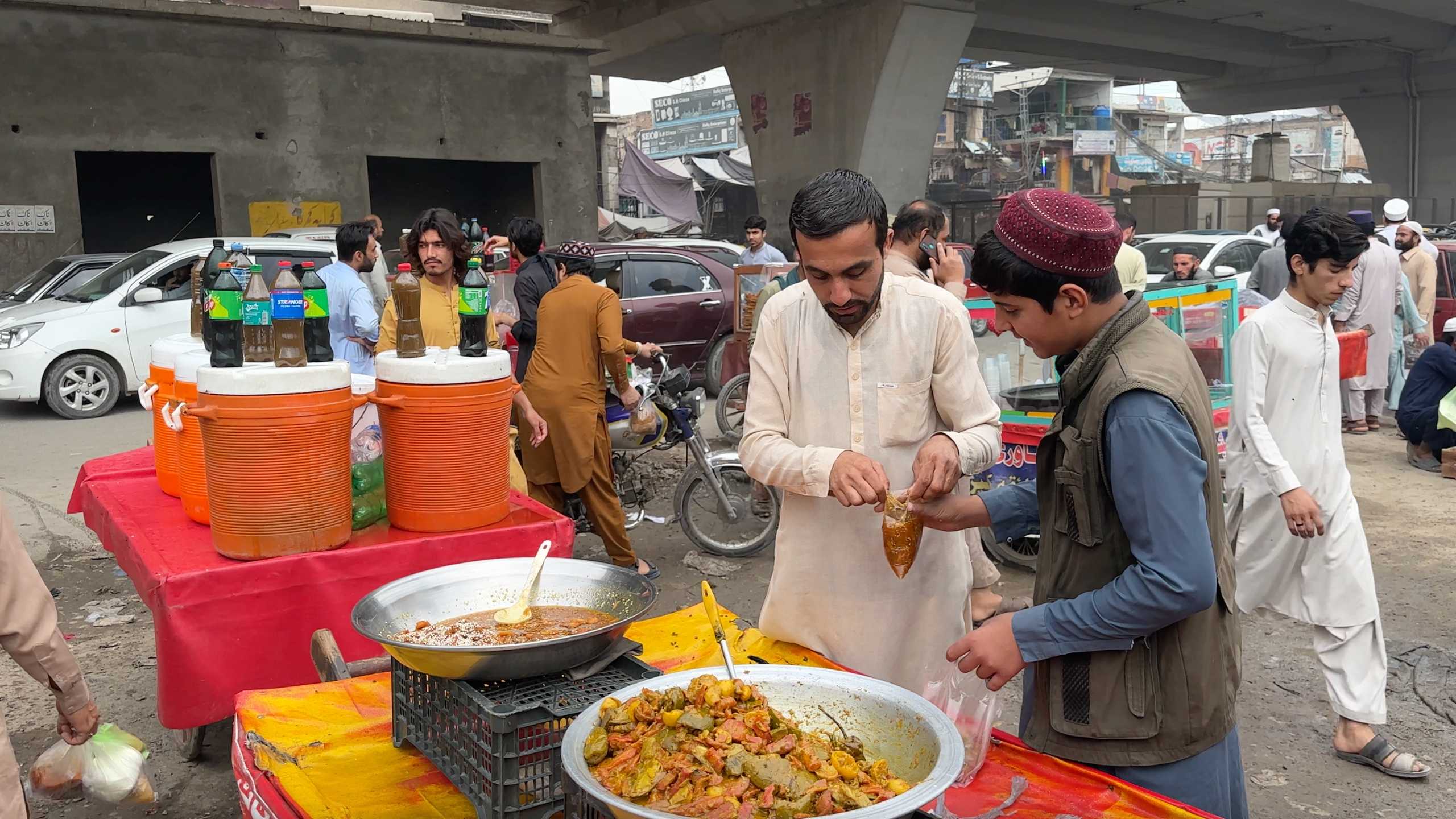 Two men serving fresh pickles at a roadside stall, surrounded by containers of cold beverages.