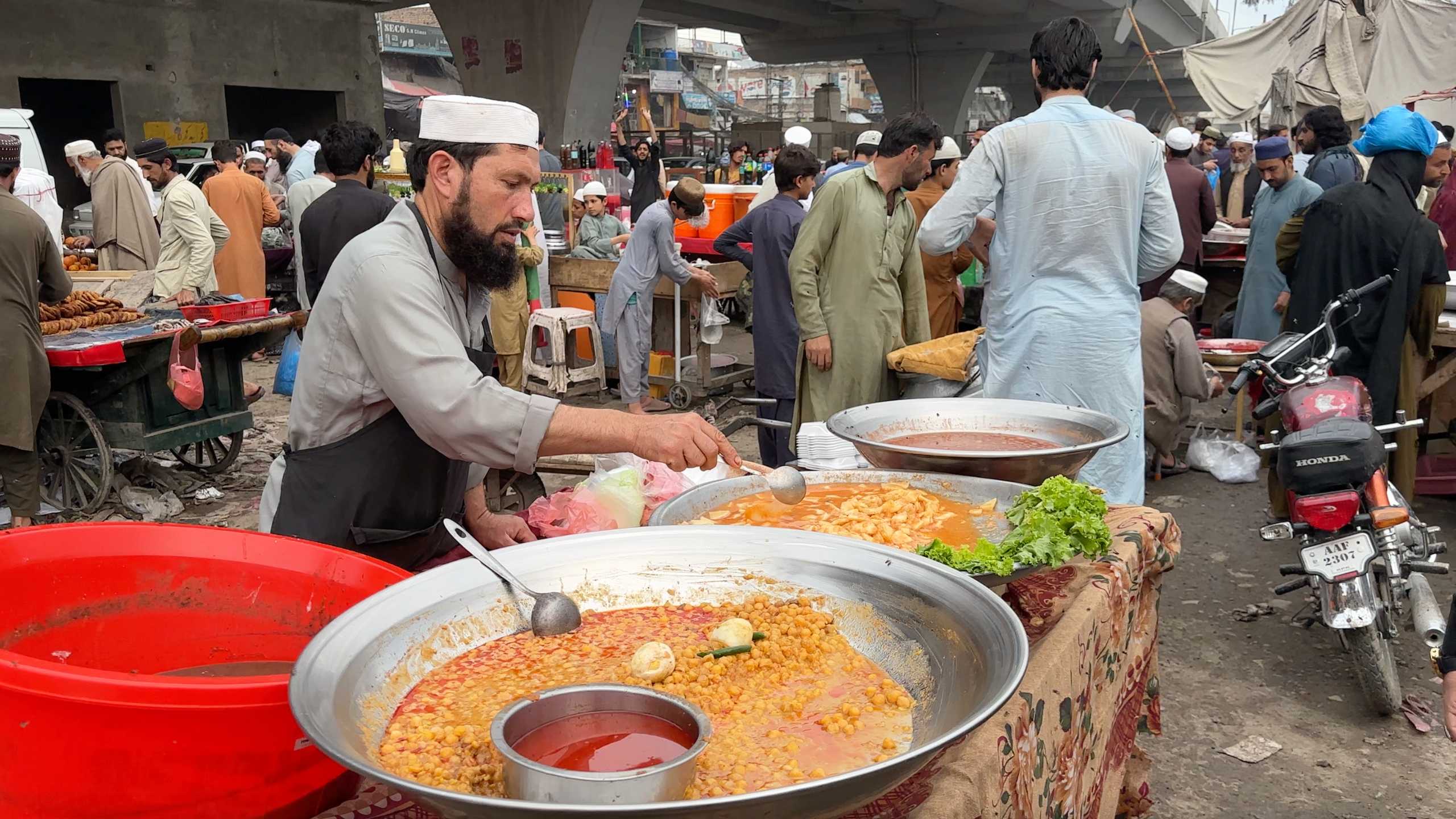 A street vendor carefully ladling out a hearty chickpea curry, with other dishes in large pots beside him.