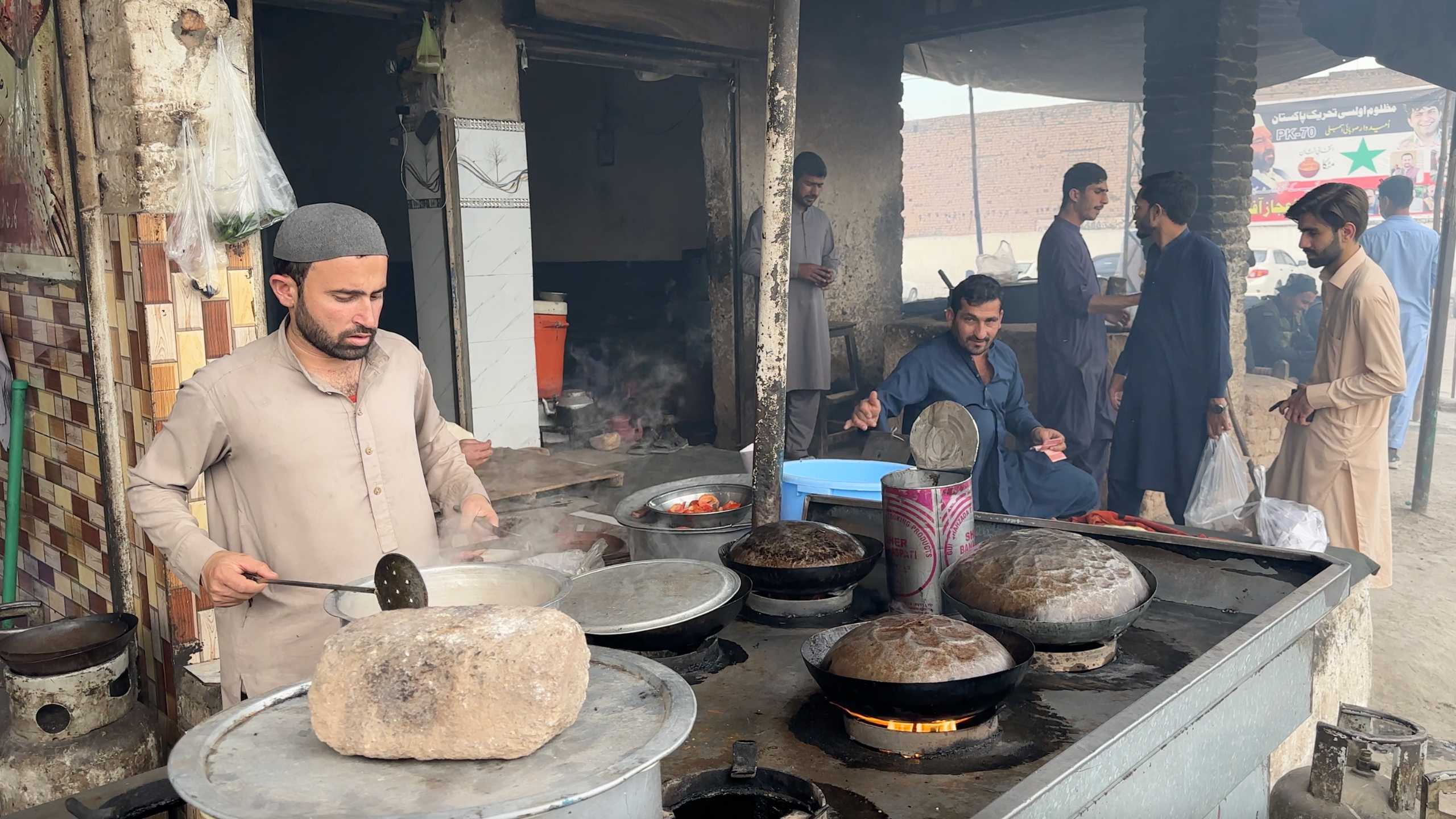 A man attentively stirs a pot in a traditional open kitchen, where flatbreads are being prepared on hot stones.