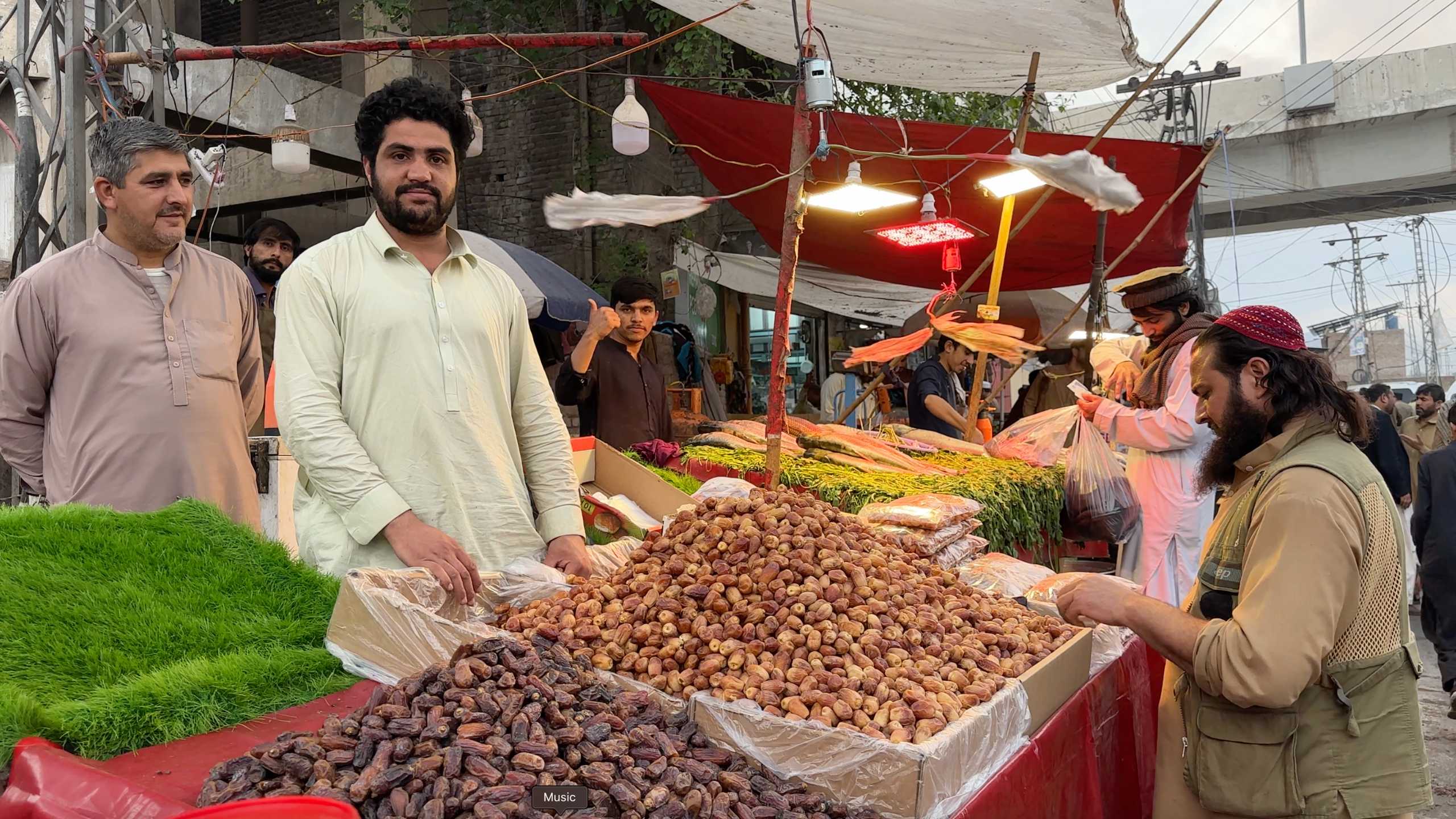 A vendor proudly displaying a large mound of dates, alongside fresh greens under a vibrant market canopy.