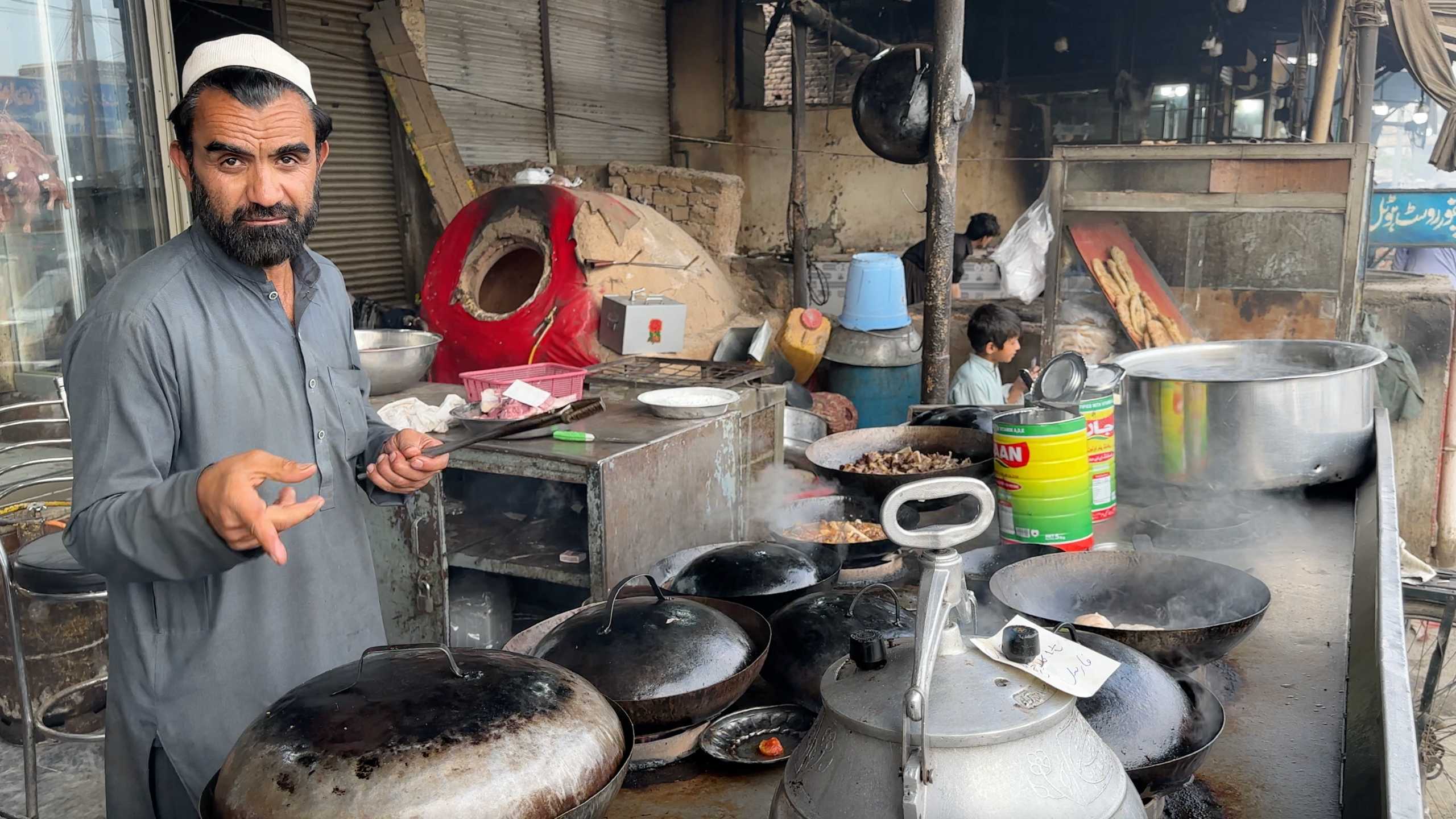 A cook stands by his makeshift kitchen, surrounded by steaming pots and a tandoor oven.