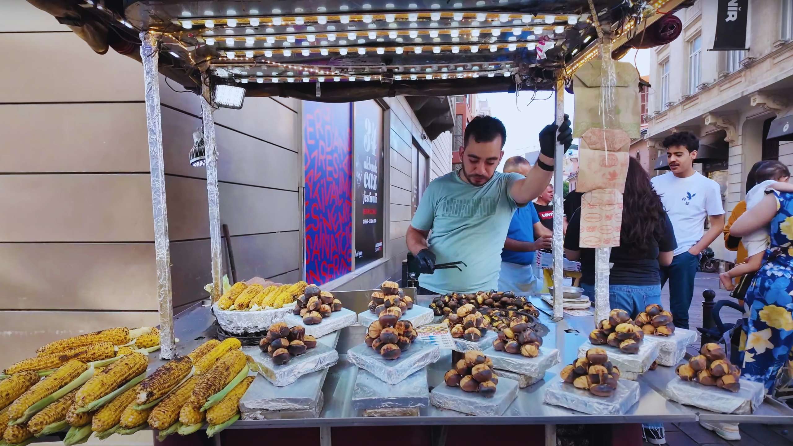 Vendor grilling corn and roasting chestnuts under string lights, attracting passersby.