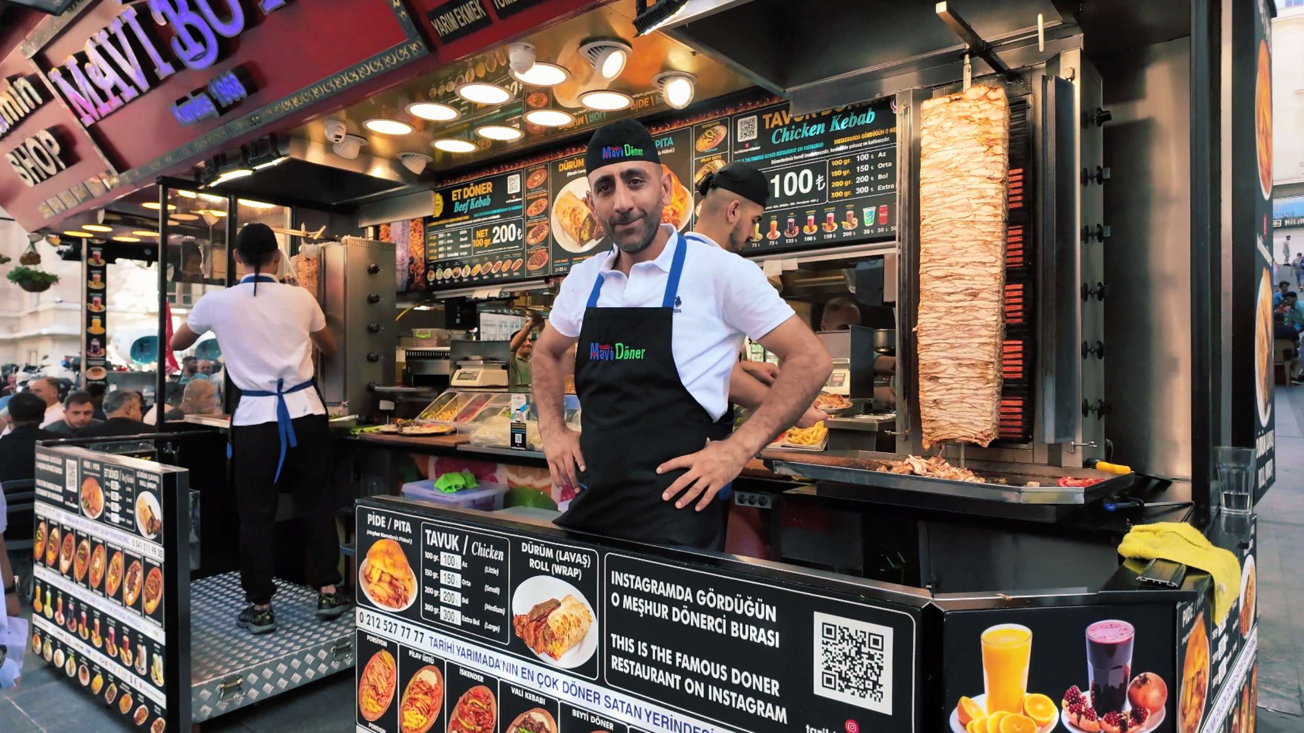 A smiling vendor stands proudly in front of a popular doner kebab stall.