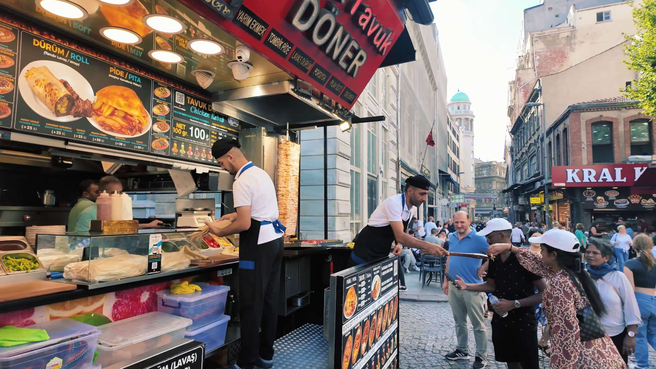 Two vendors prepare and serve doner kebabs while chatting with customers.