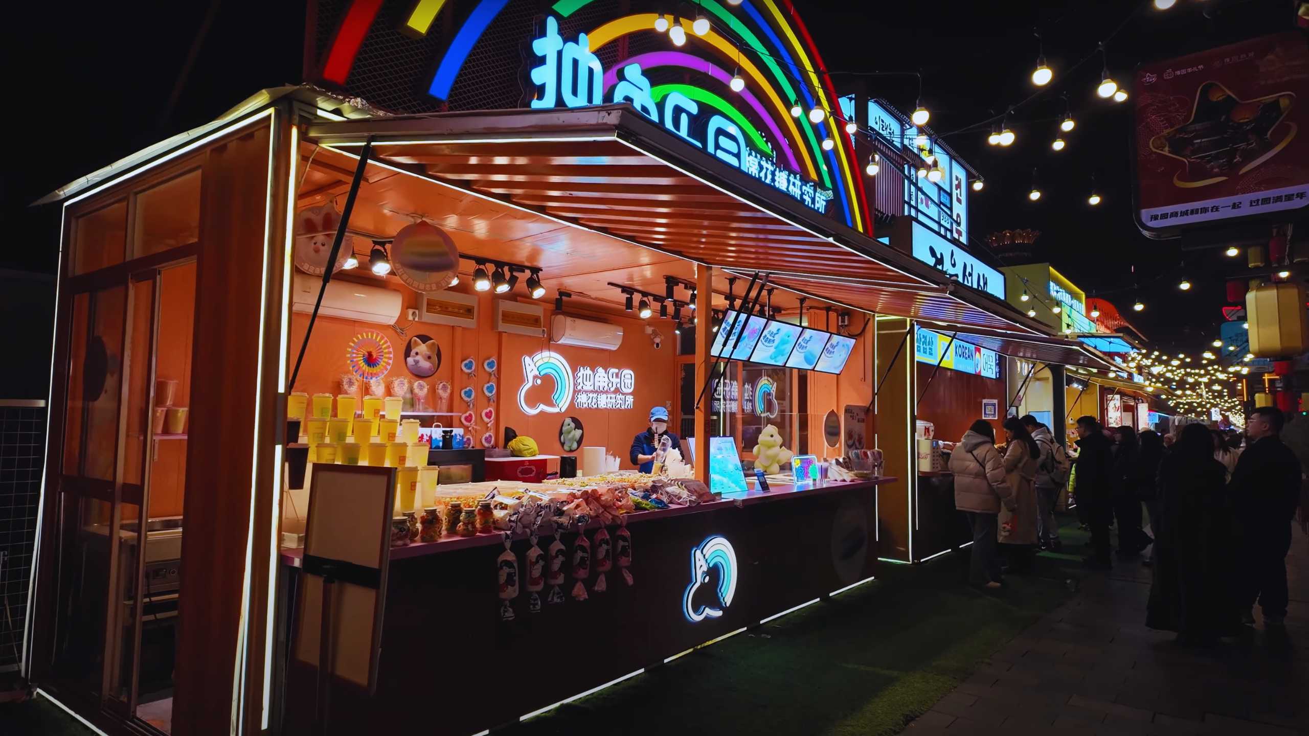 Vibrant food stall with rainbow signage illuminating the night.