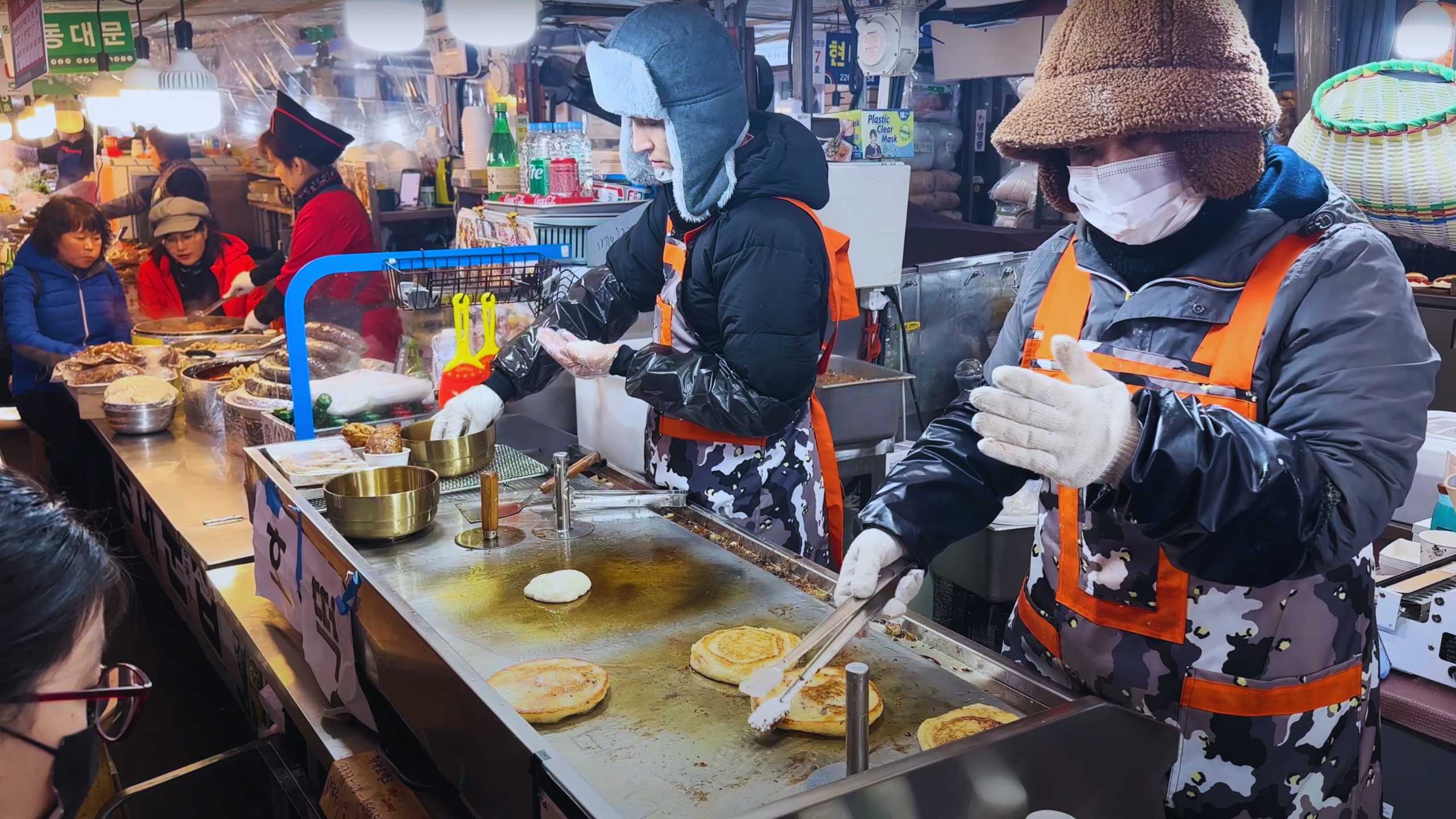 Vendors in warm clothing preparing pancakes on a griddle, while customers enjoy the bustling food scene.