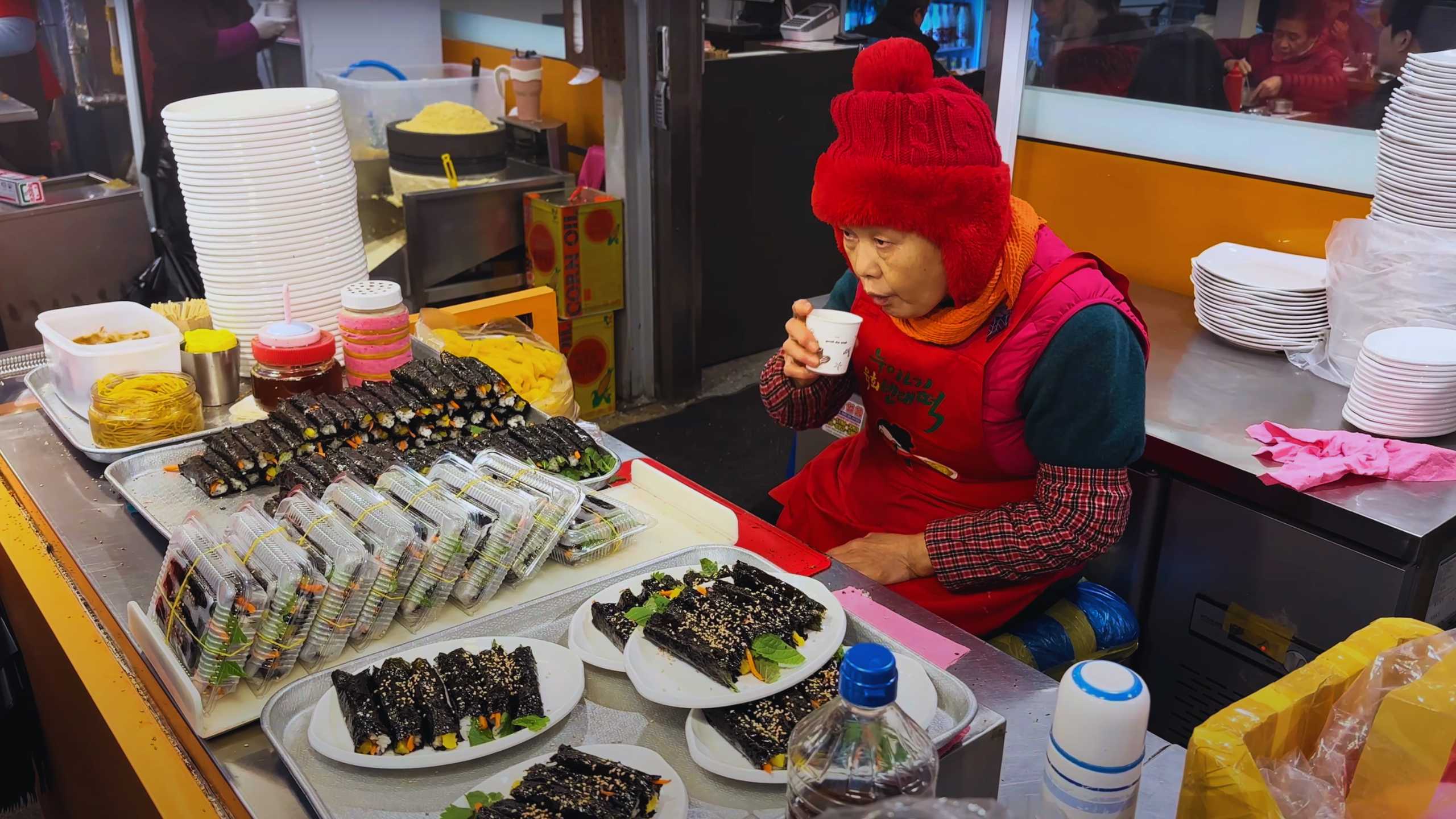 A vendor taking a quick break with a drink, surrounded by neatly arranged rolls of gimbap.