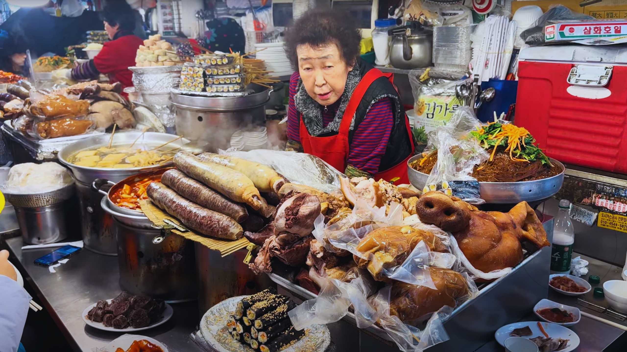 An older woman overseeing a table laden with traditional Korean dishes, including sundae and pig's head.