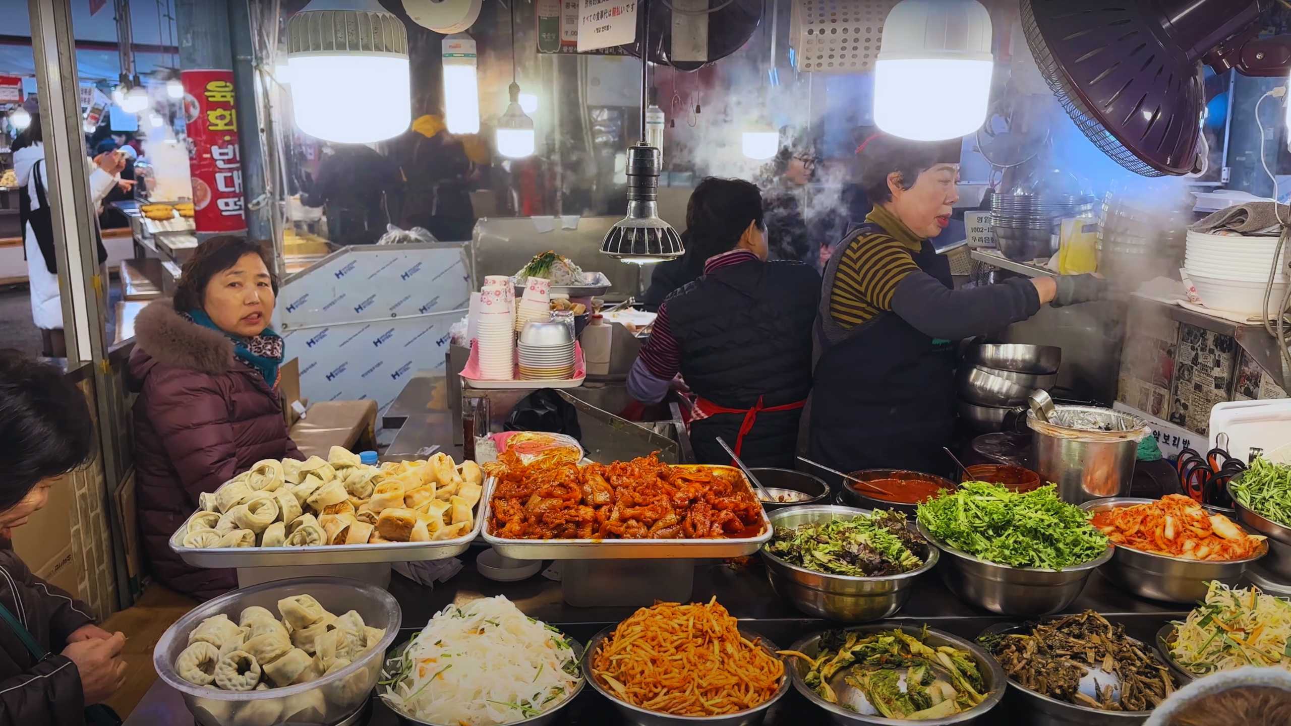 A vendor prepares a variety of Korean dishes, with dumplings, noodles, and vegetables on display, while customers await their meal.