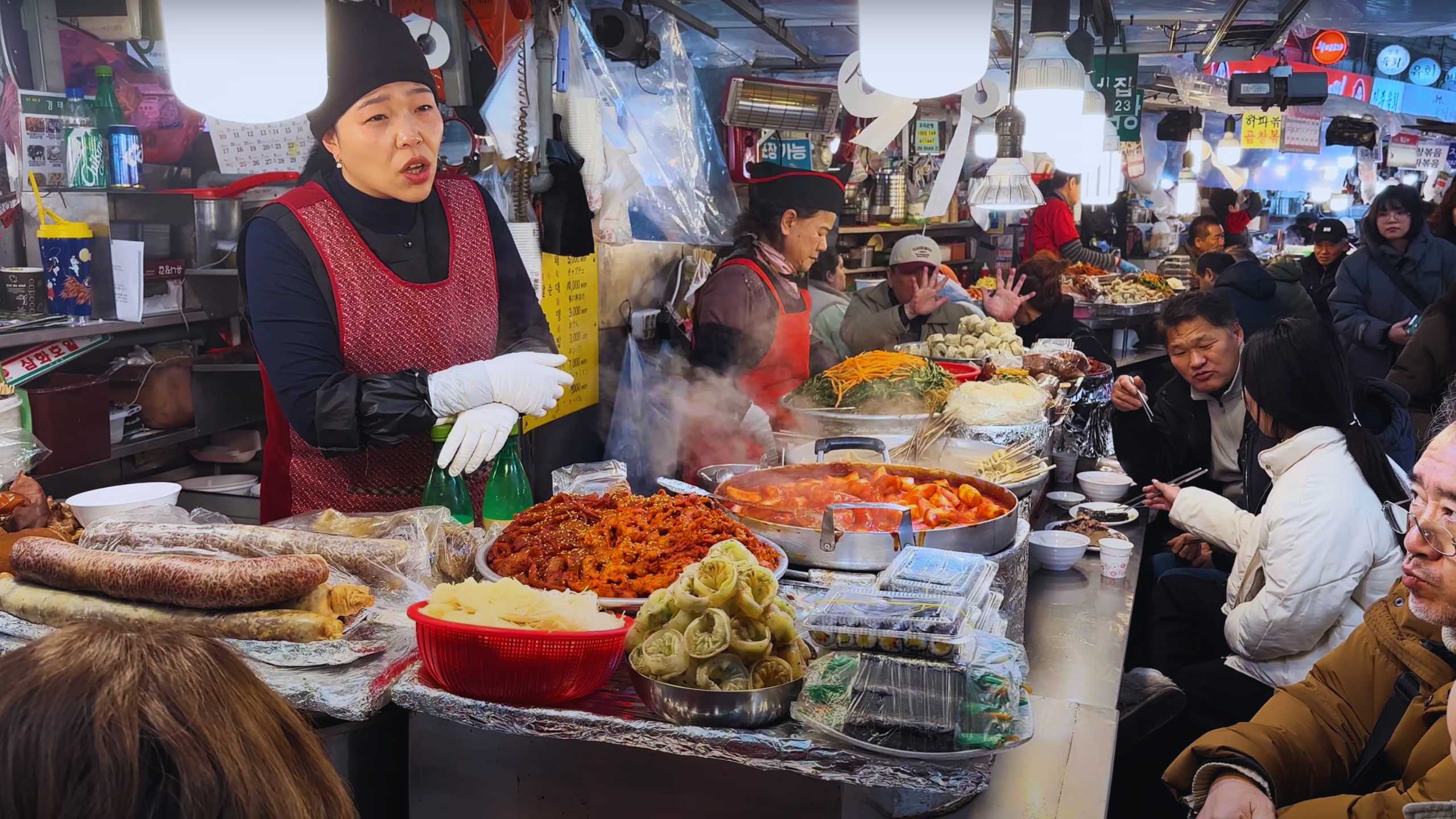 A bustling food stall offers steaming dishes, including tteokbokki and kimbap, as a vendor converses with customers.