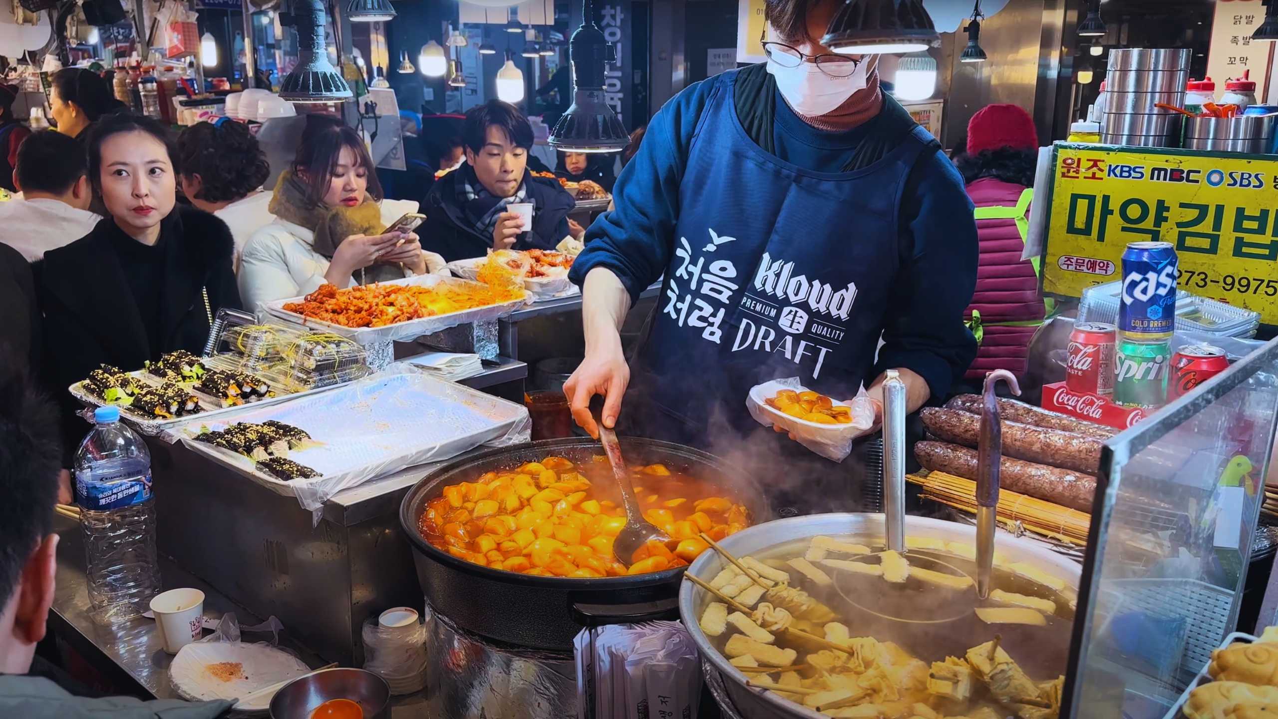 A vendor stirs a large pot of tteokbokki, while nearby, rolls of gimbap await hungry customers.