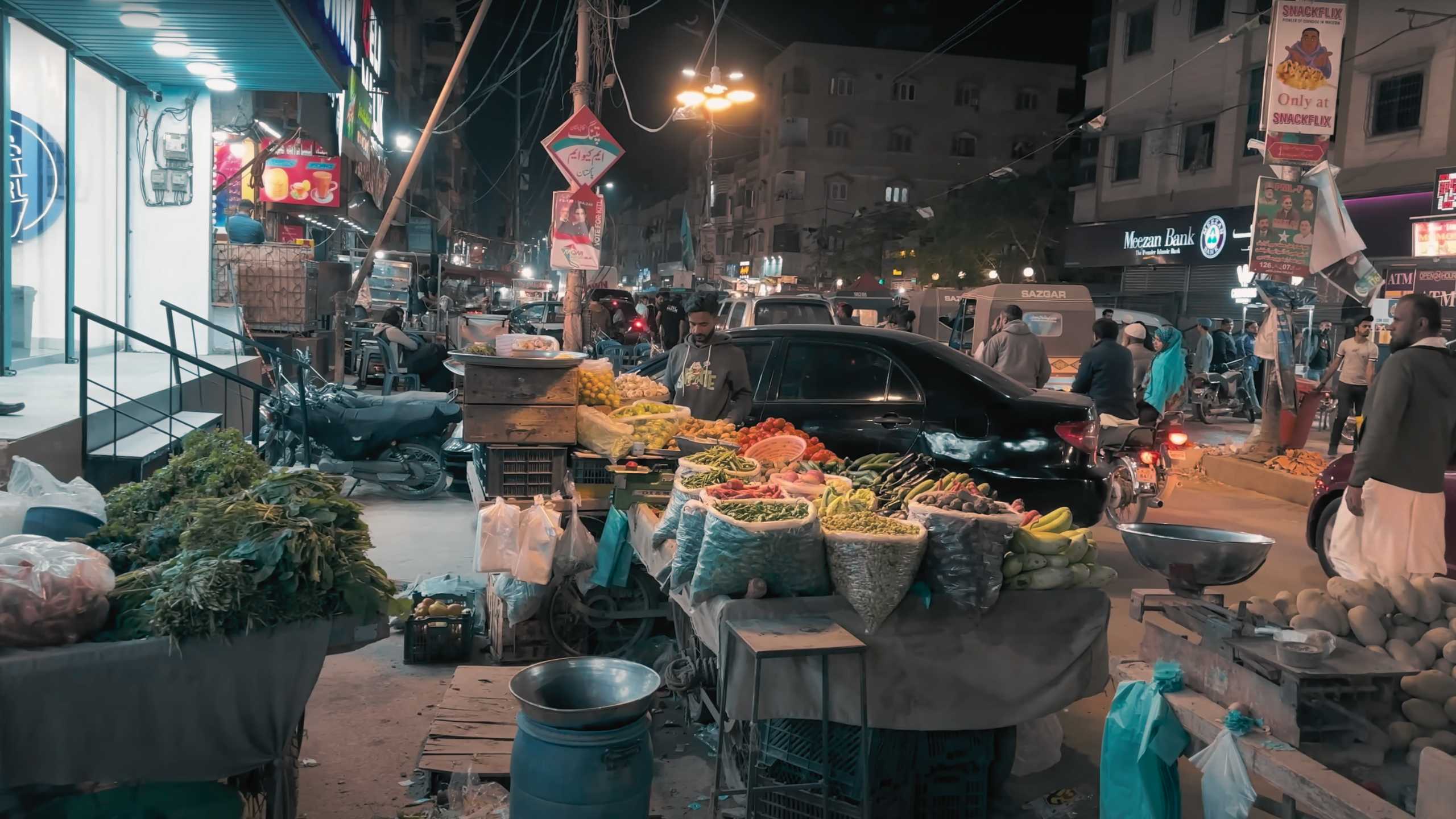 A busy night market scene with a vegetable vendor offering fresh produce under glowing streetlights.