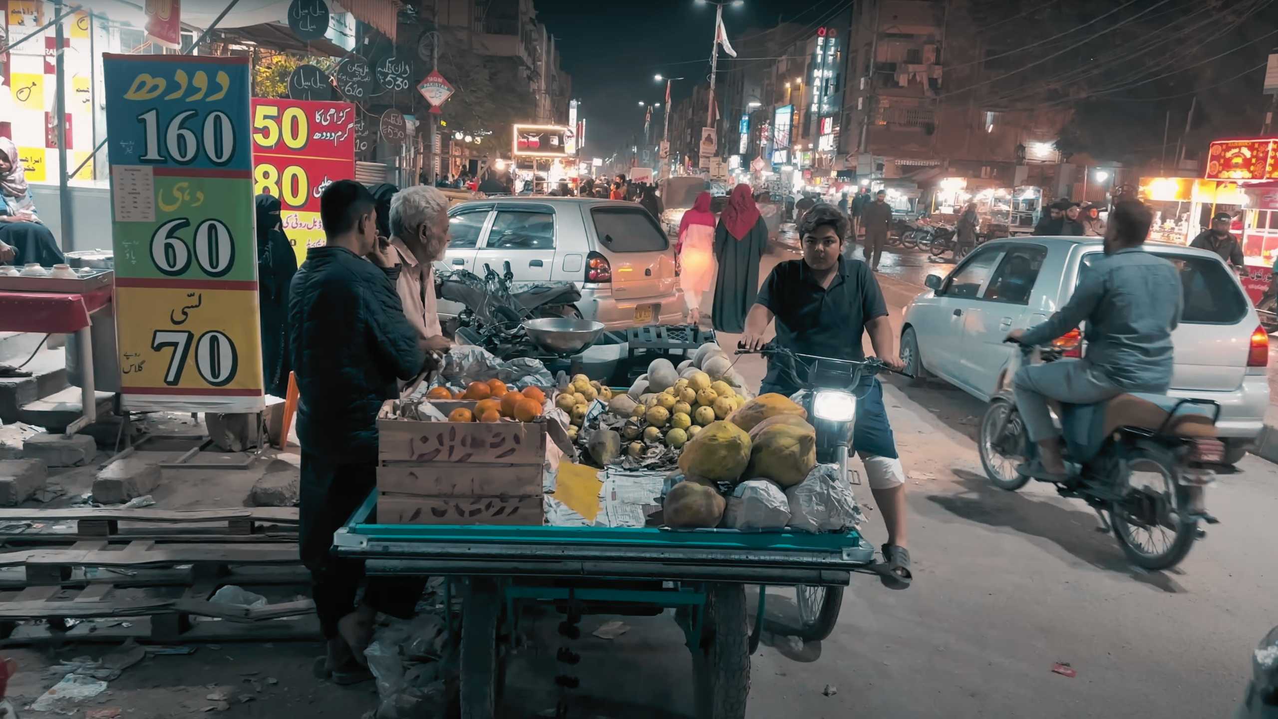 A street vendor selling fresh fruits under colorful signs as motorcycles and cars pass by.