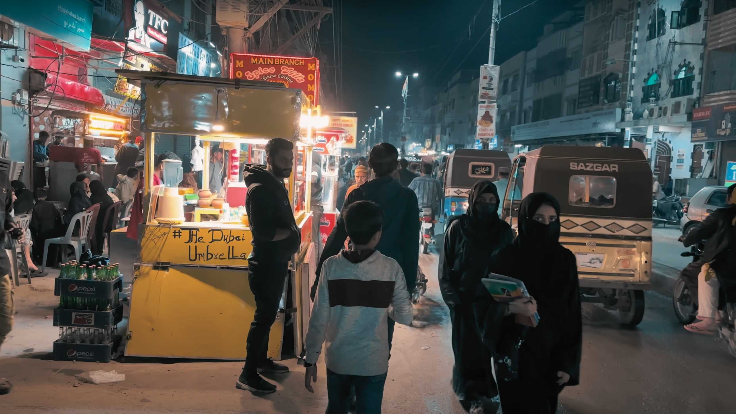 Families stroll along a busy street, passing by brightly lit food stalls.