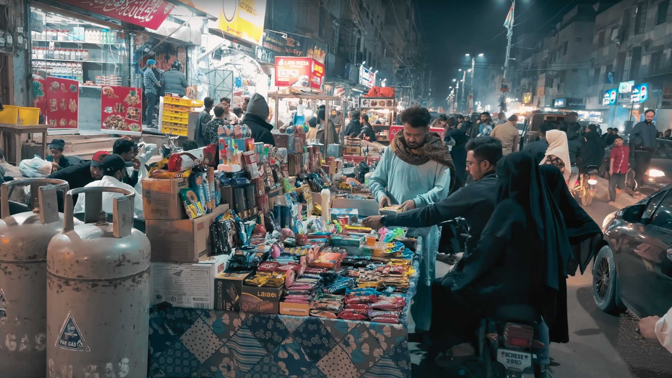 A vendor sells assorted snacks and candies as pedestrians browse the busy market.