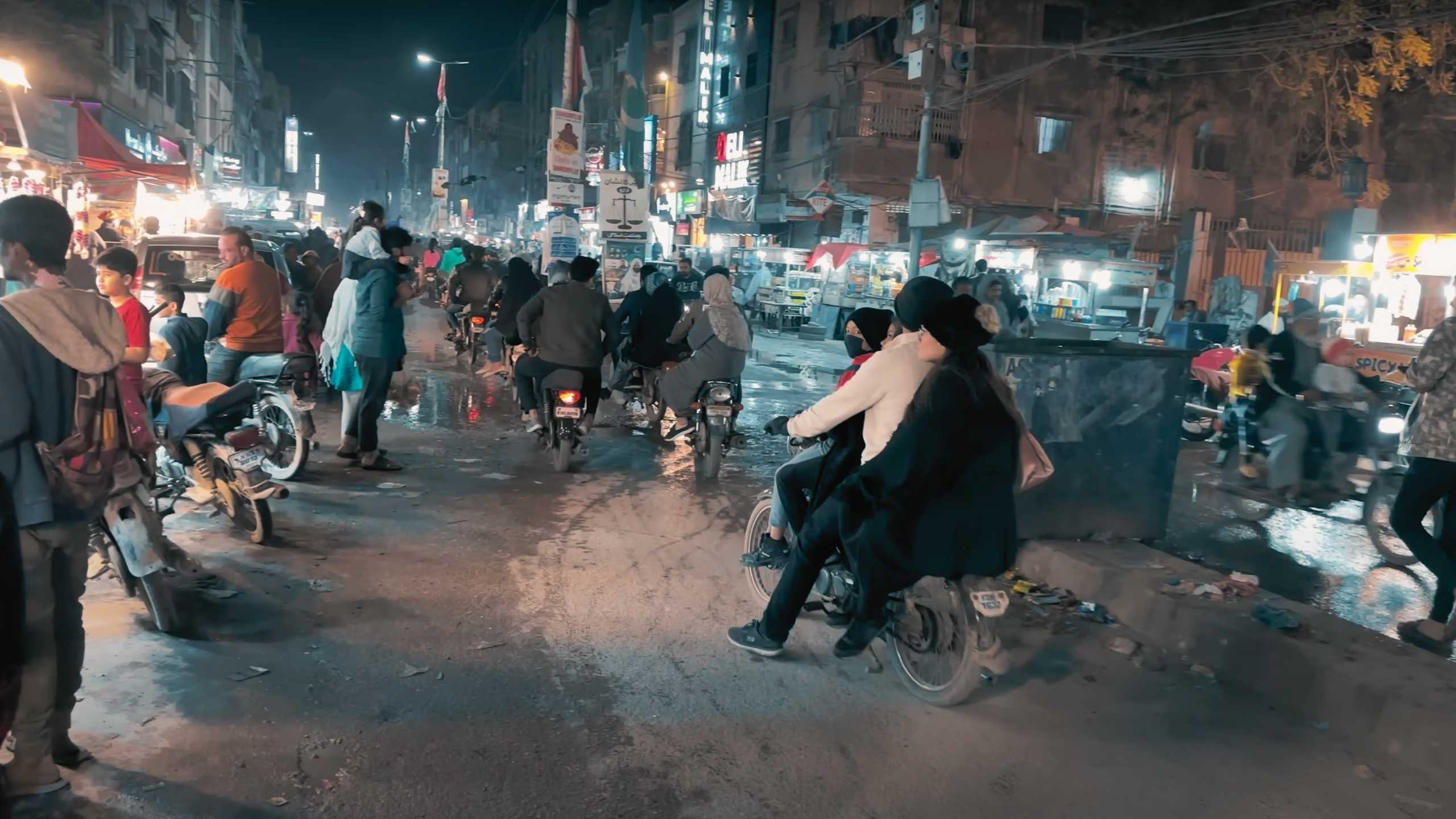 Motorbikes navigate through a crowded, lively street, adding to the energetic atmosphere.