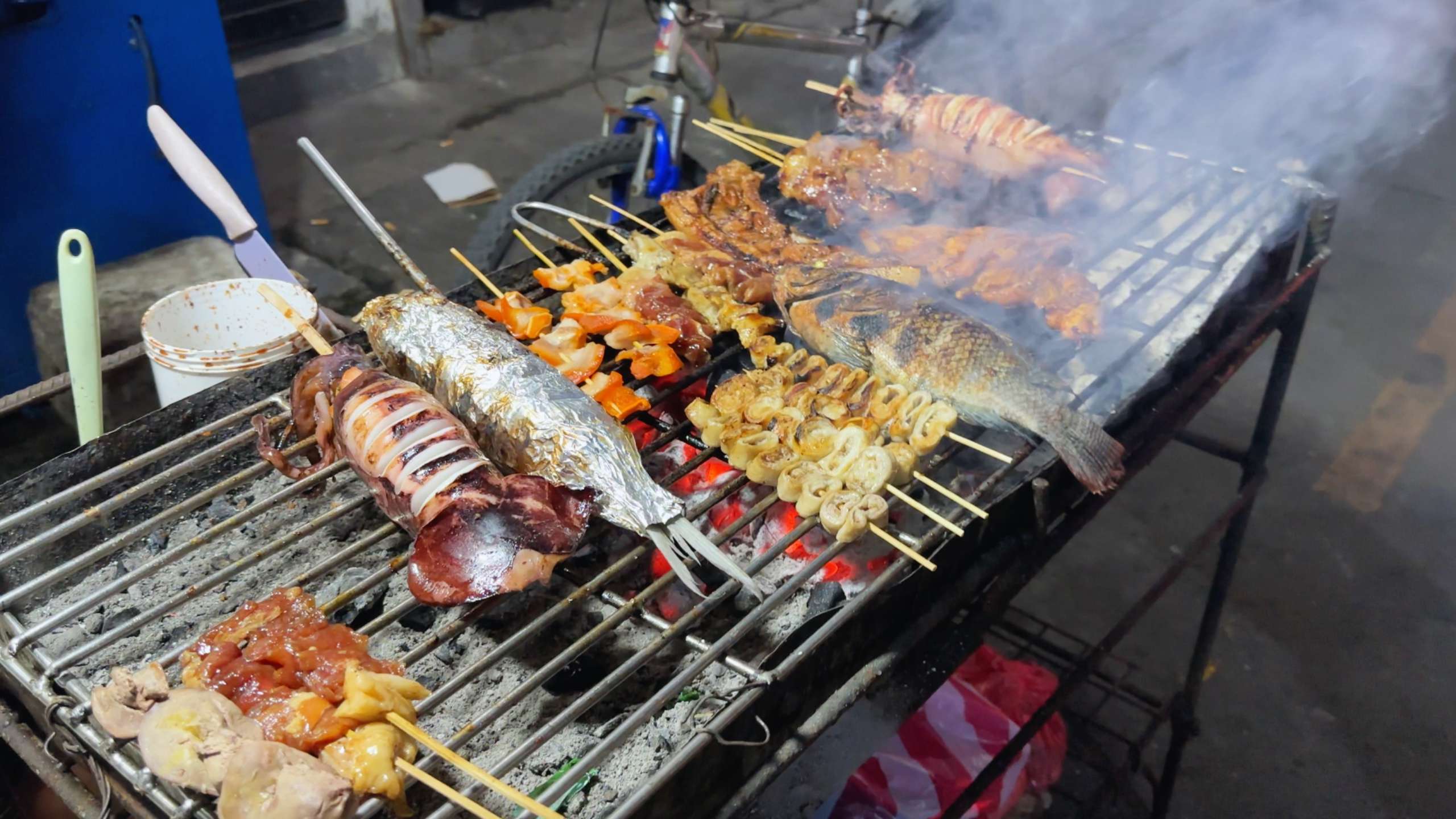 A variety of skewered seafood and meats sizzling on a charcoal grill, filling the air with smoky aromas.