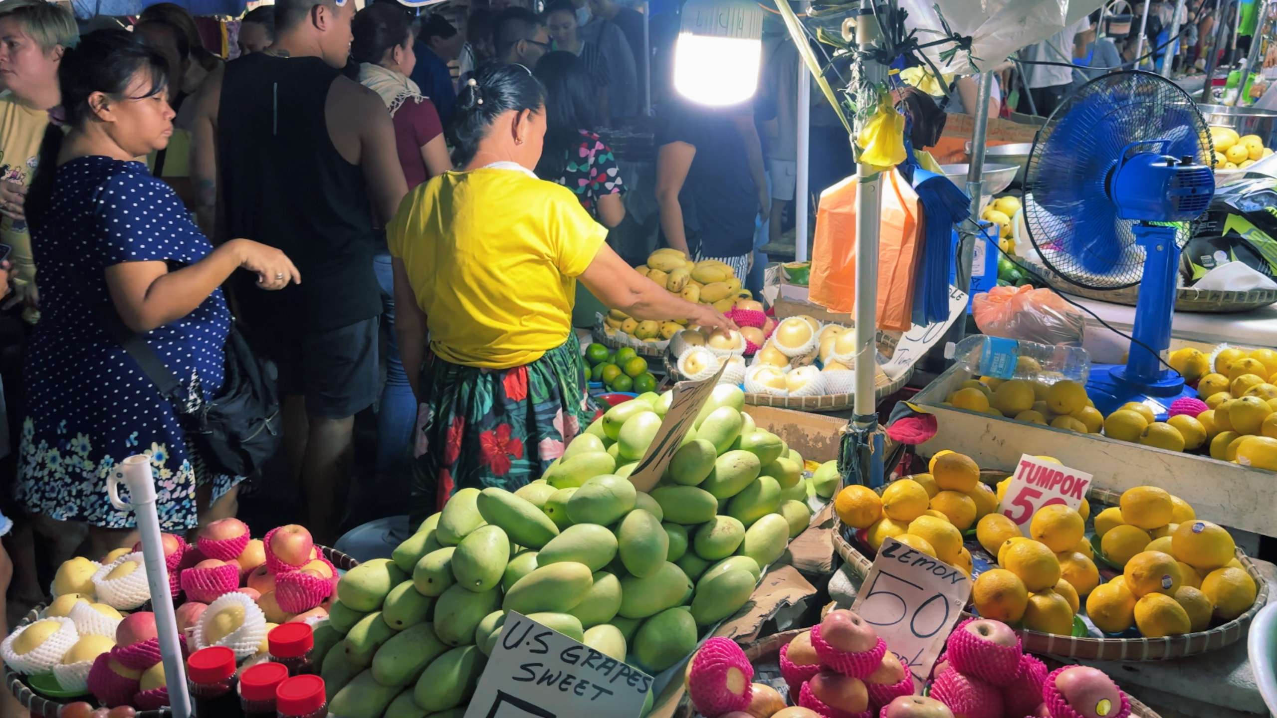 A woman sells various fruits, including mangoes and apples, in a vibrant, colorful stall.