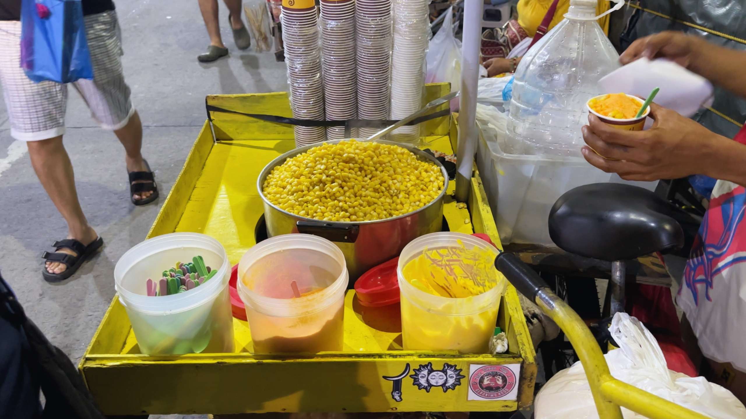 A vendor serves boiled corn with butter, seasoning options, and colorful spoons.