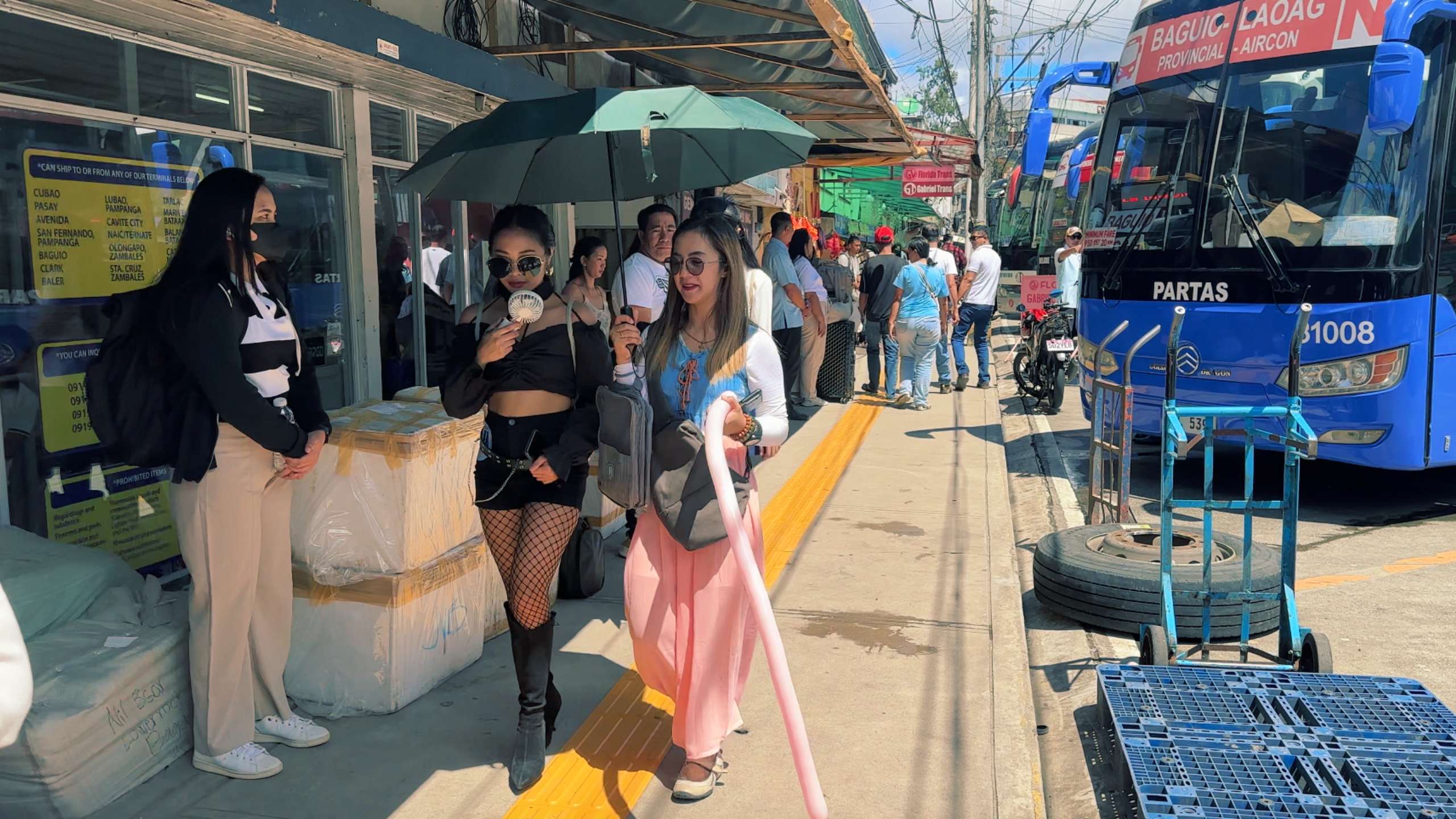 Two stylish women walk under an umbrella, enjoying cold drinks on a sunny day.