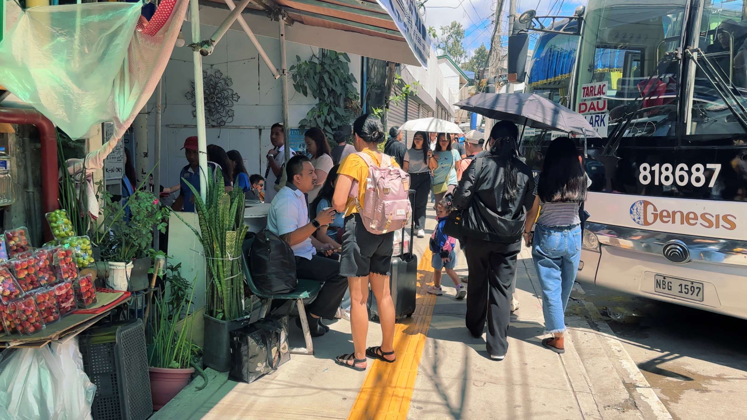 Crowds gather near a bus station, with vibrant street stalls and umbrellas for shade.