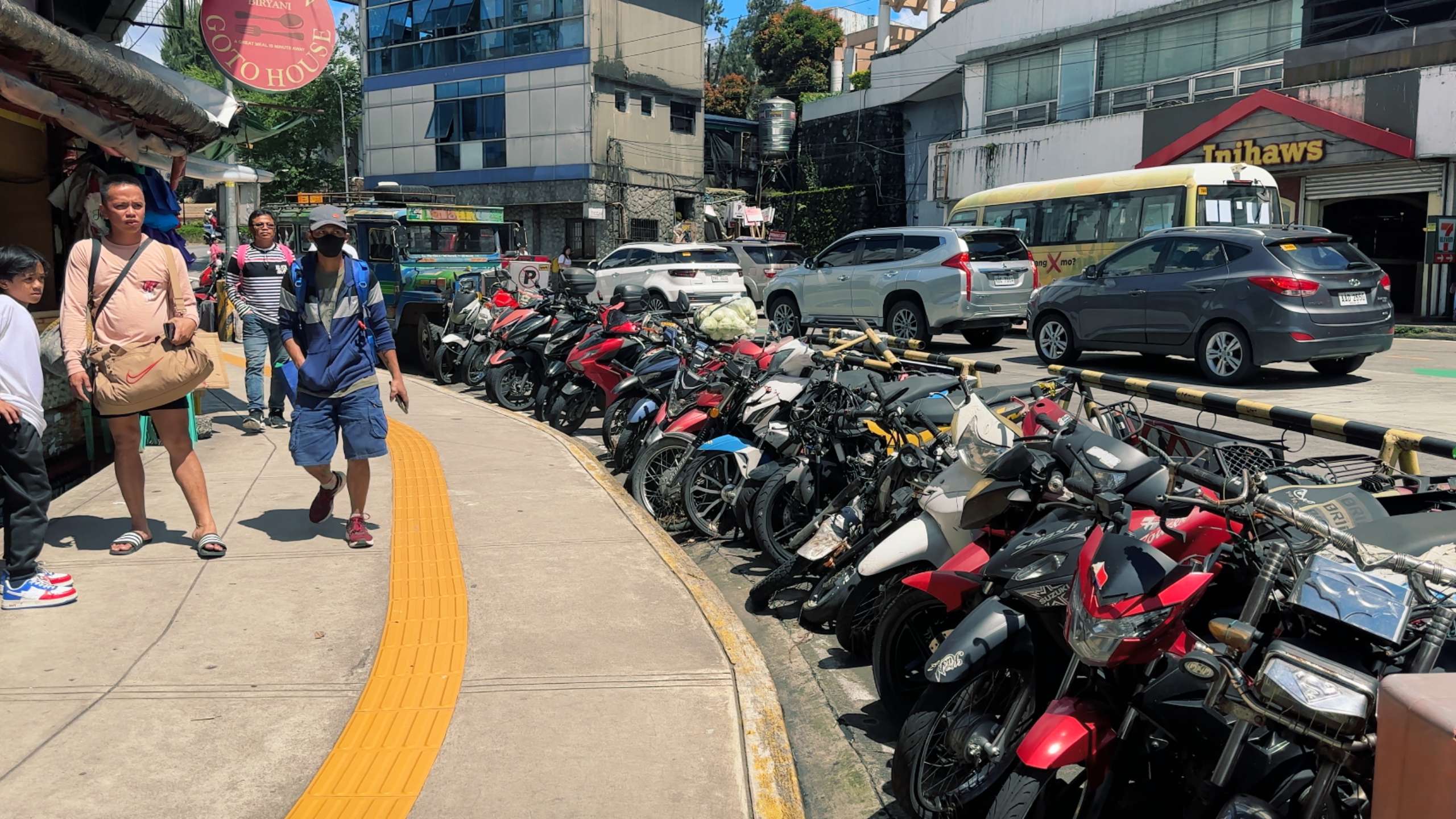 A row of parked motorbikes lines a busy sidewalk in a bustling urban area.