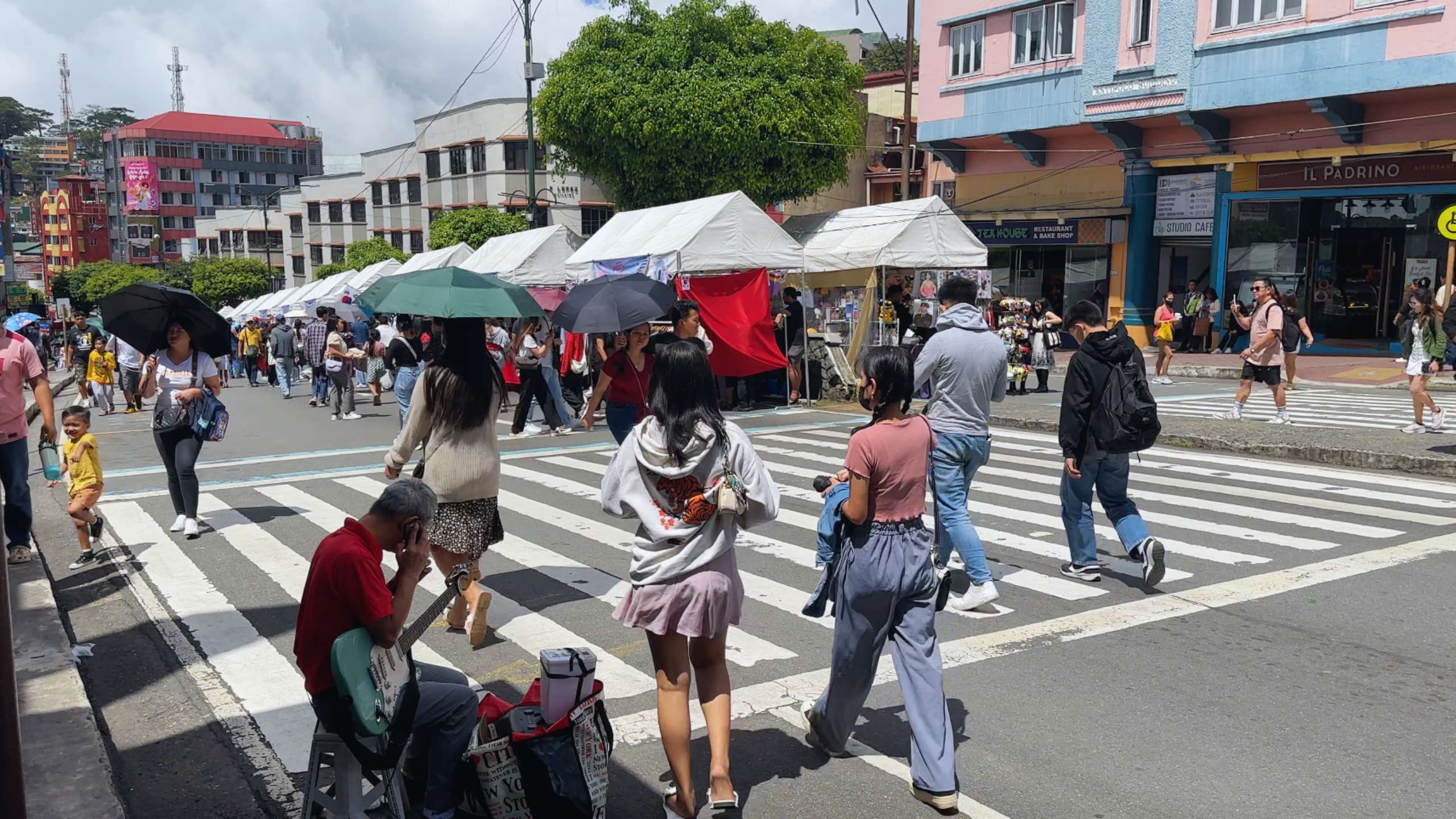 Pedestrians cross a busy street lined with white tents and shaded vendors.